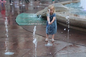 Una niña se lo pasa de miedo correteando entre los chorros y cogiendo agua de unos y de otrso